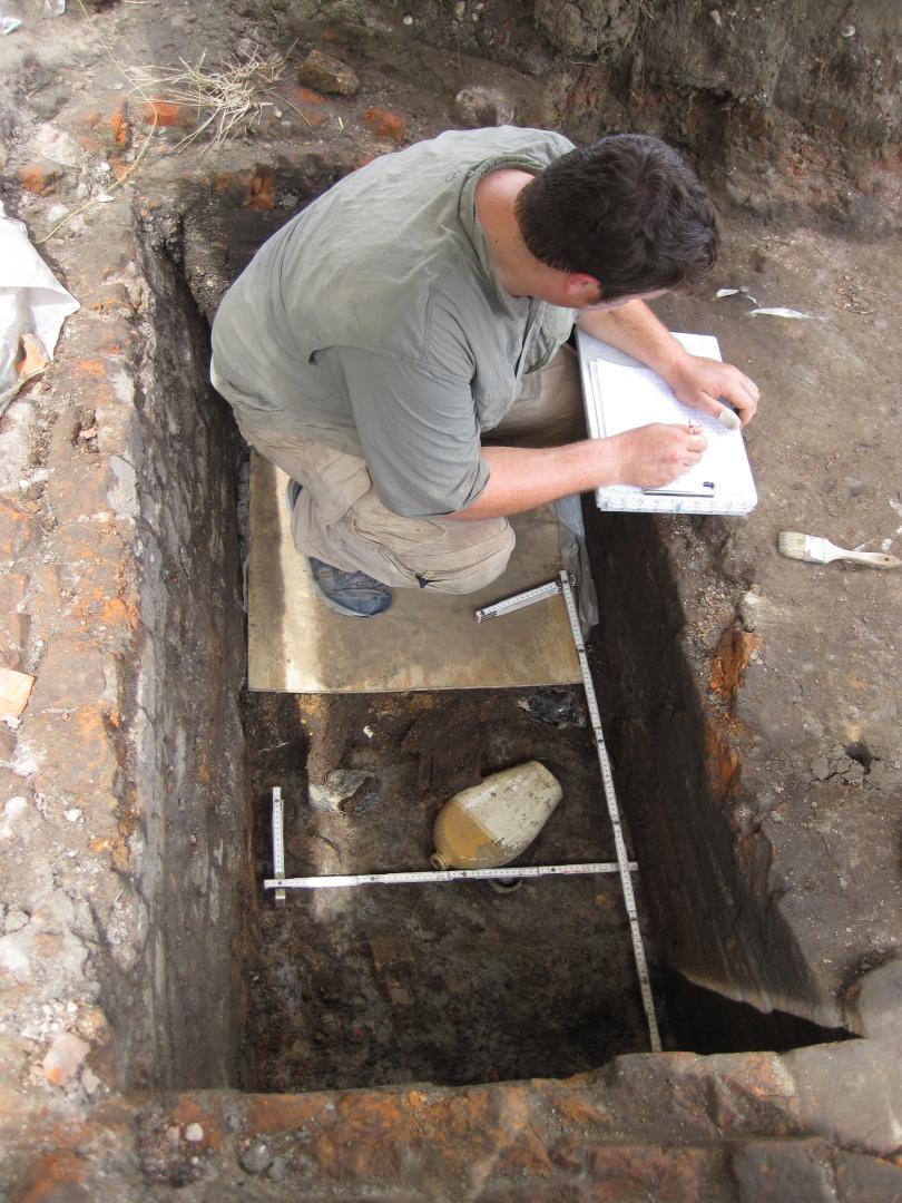 Image of two archaeologists recording and excavating a test unit with historic New Orleans homes in the background