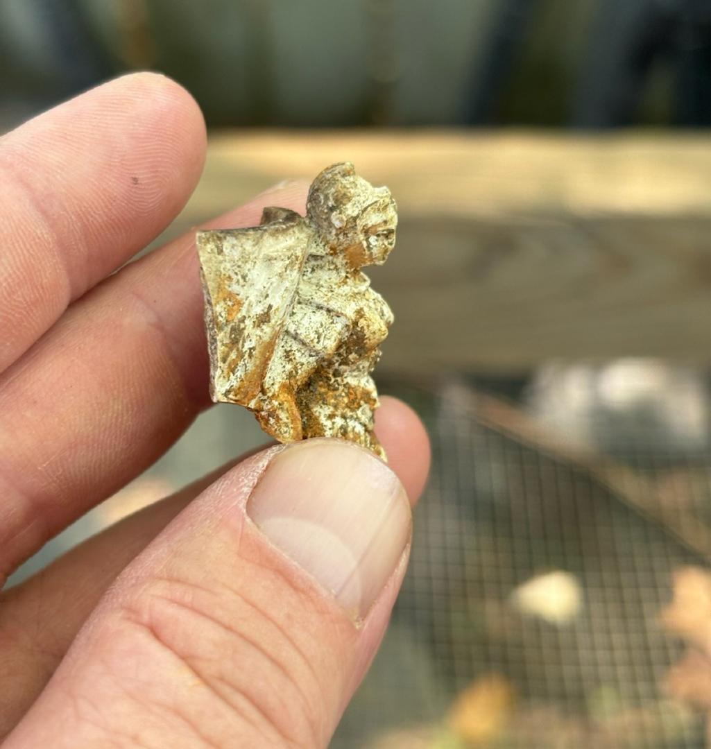 Photograph of a pipe molded in the image of a woman with a helmet, held between the fingers of an archaeologist.