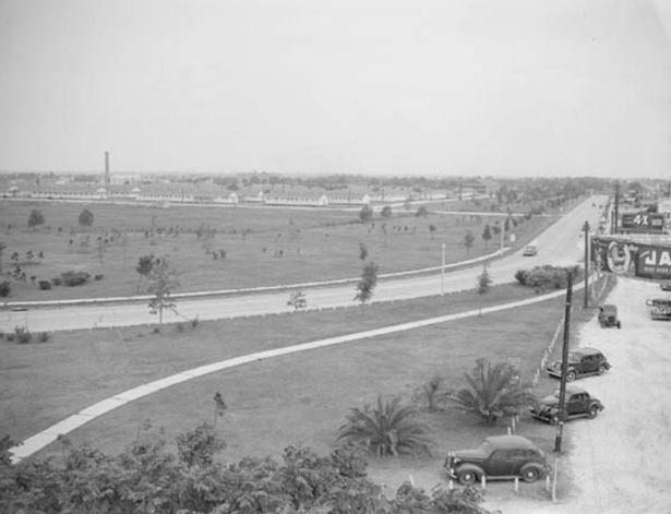 Black and white photo showing a road extending off into the distance, with a few vintage cars parked in the foreground. In the distance there are several long, white buildings.