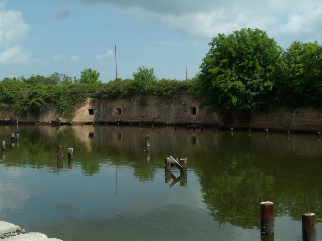 Color photograph of a brick fort surrounded by water with greenery growing on top of the fort and wooden pilings sticking out of the water in the foreground.