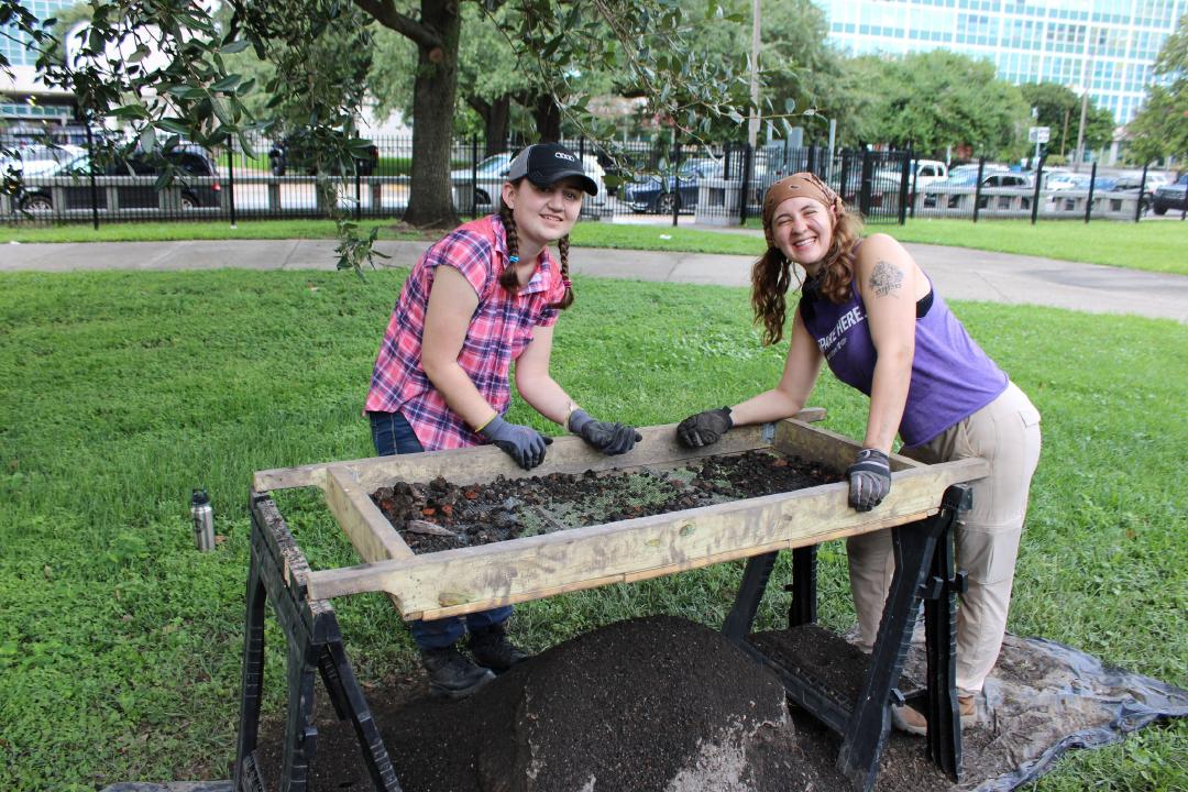 Photograph of two students around a wooden rectangular screen frame propped up on saw horses. A mound of dirt has been sifted through the quarter inch mesh screen.