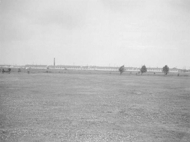 Black and white photo. An expansive lawn stretches out to a sparse line of trees. Beyond that, a long low white building in the distance.