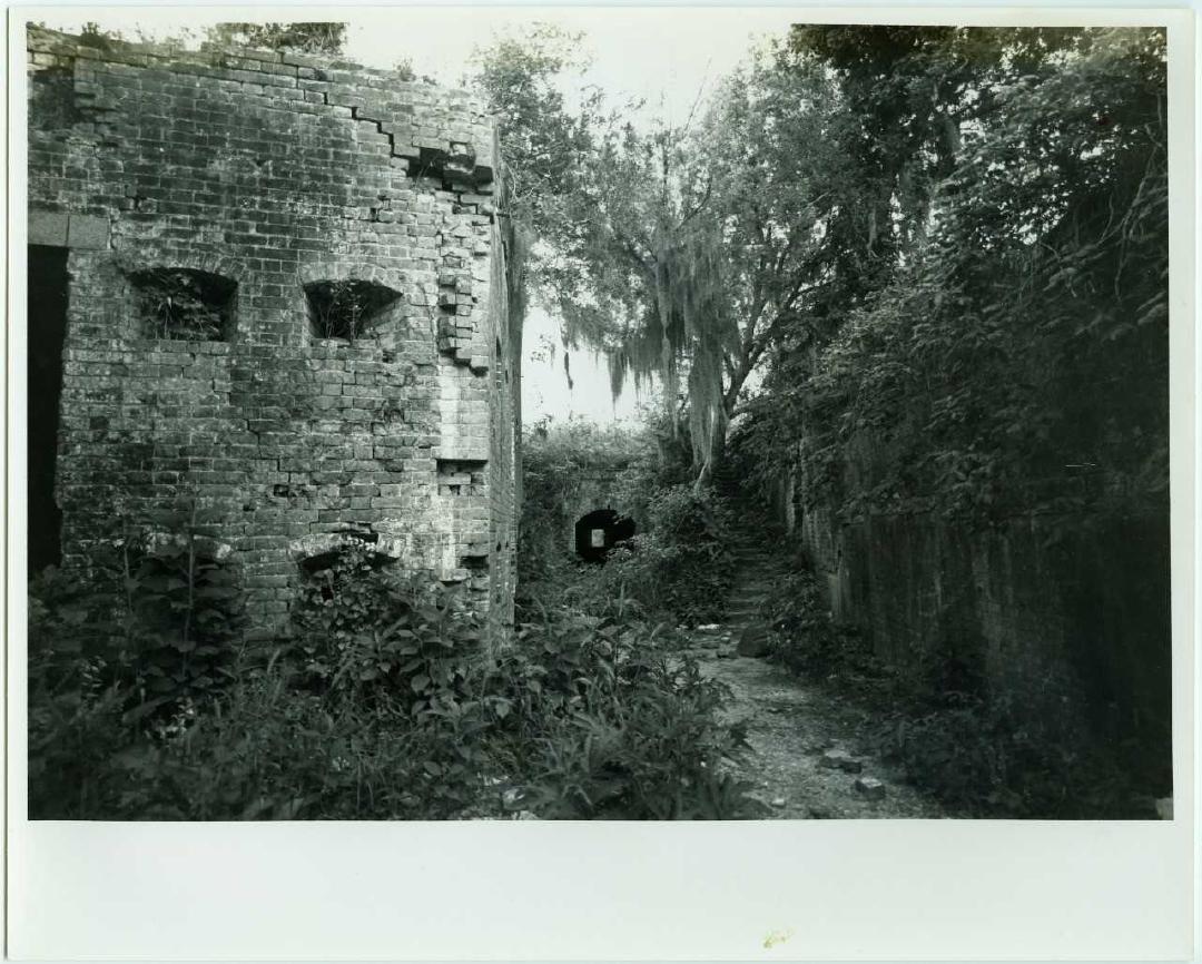 Black and white photo of ruin of brick fort overgrown with greenery and with fallen bricks visible on the ground.