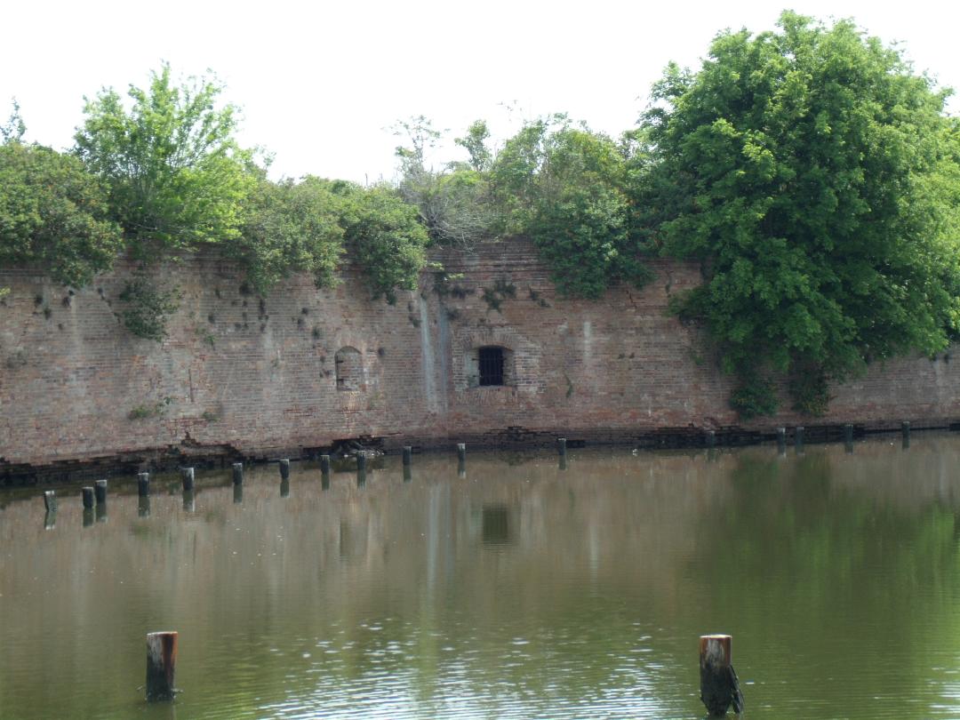 Color photograph of a brick fort surrounded by water with greenery growing on top of the fort.