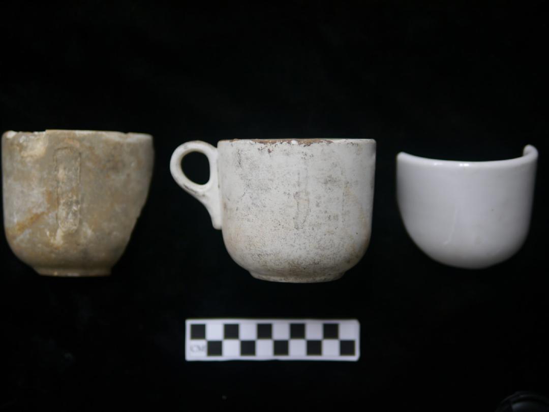 A photograph of three cup fragments, all similar shape and size. On the right, a brown and white clay biscuit coffee cup without handle; center, a white clay biscuit coffee cup with handle; left, white glazed coffee cup fragment. Photograph is on black velvet with black and white centimeter bar below the pottery fragments.