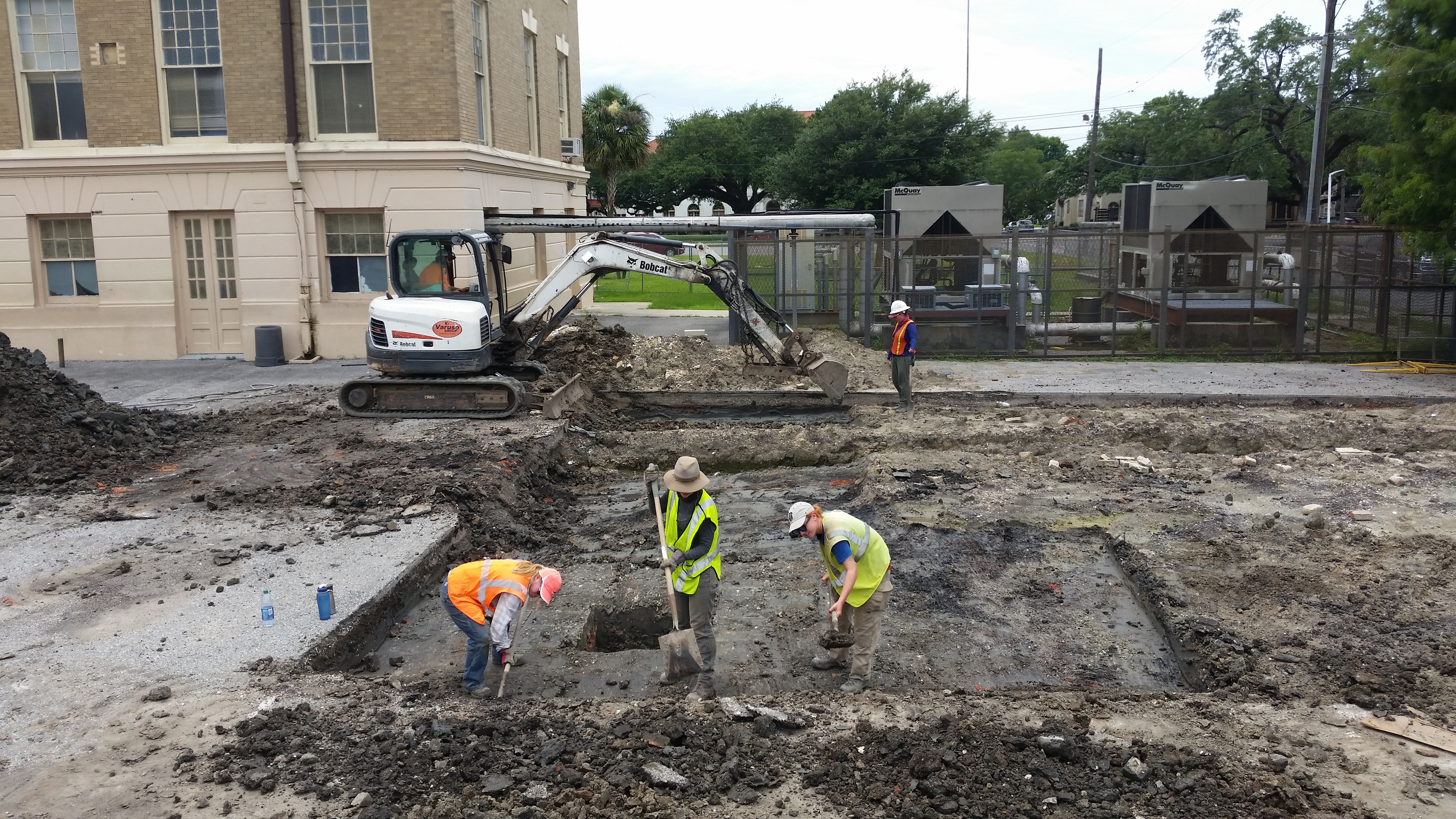 Three people in safety equipment in the foreground using shovels to clear off top layer of soil. Backhoe digging in the background, just in front of the school building