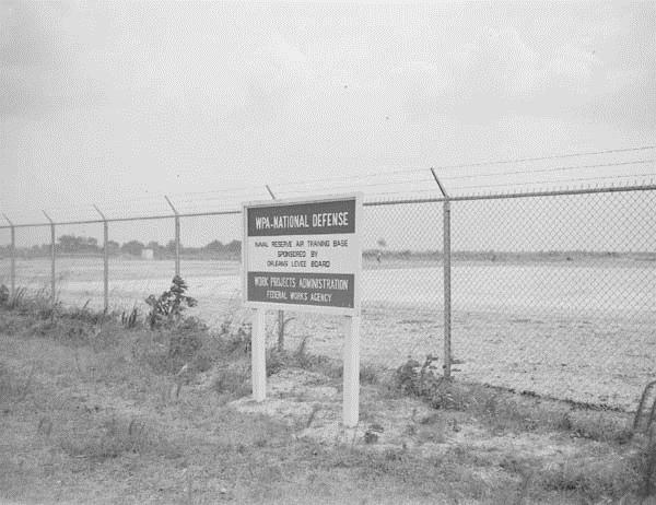 A sign in front of a chain link fence topped with barbed wire. The sign heading reads "WPA- National Defense"