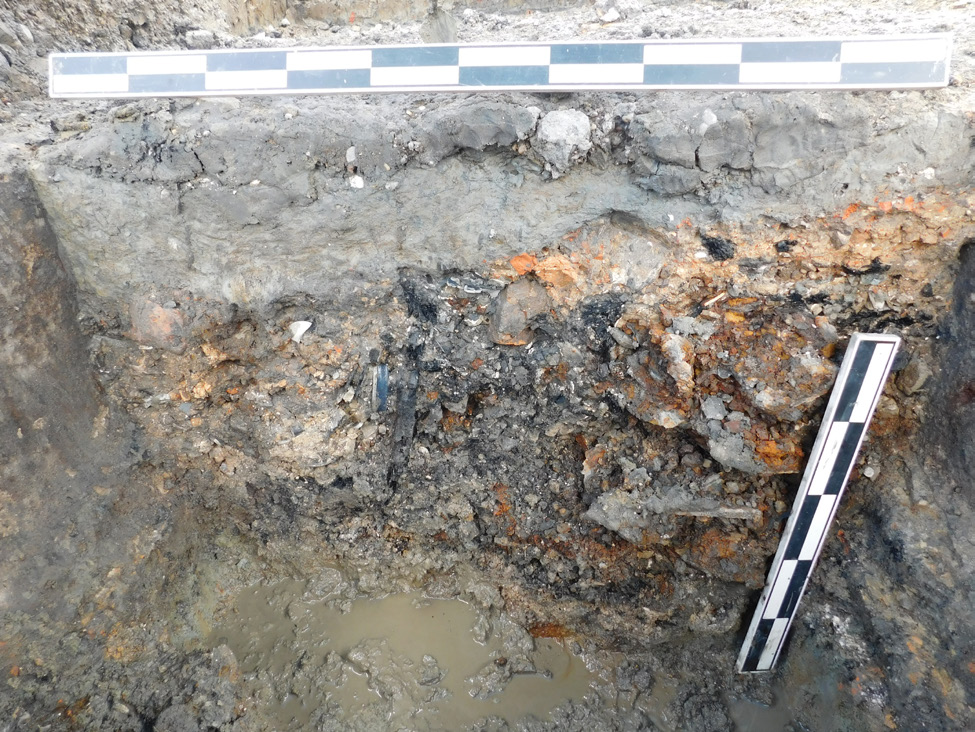 Side wall in square excavation unit with pile of charred and rusted rubble to the right