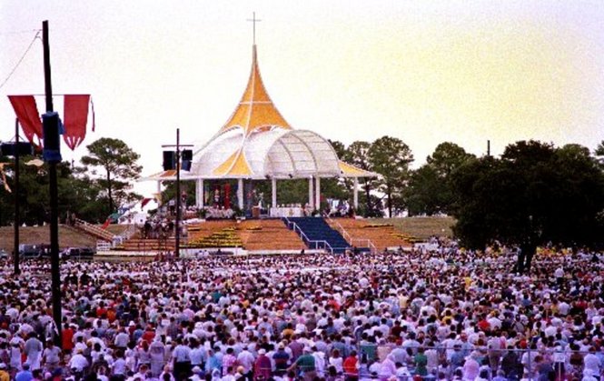 A large crowd is assembled around a raised platform with a pointed white and yellow shelter over it. There is a cross at the top of the point.