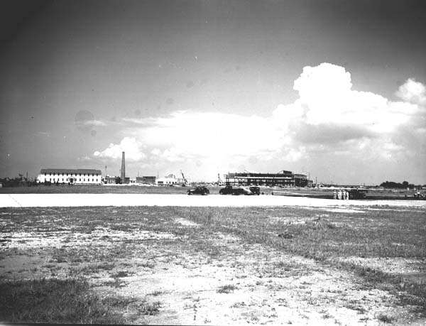 Black and white photo of open field with under construction building and smokestack in the background.